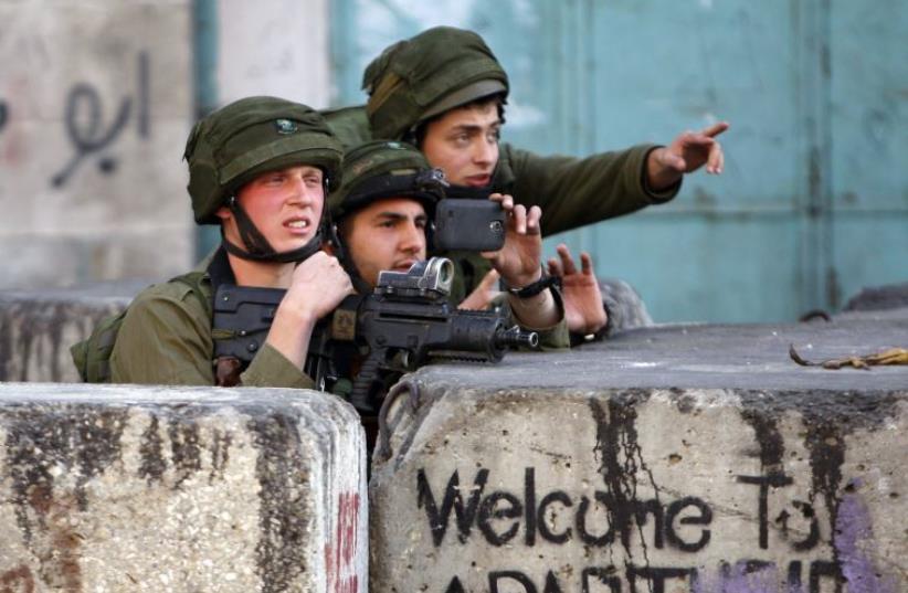 An Israeli soldier uses his mobile to take pictures during clashes with Palestinian stone-throwers in the West Bank city of Hebron March 6, 2015 (photo credit: MUSSA QAWASMA / REUTERS)