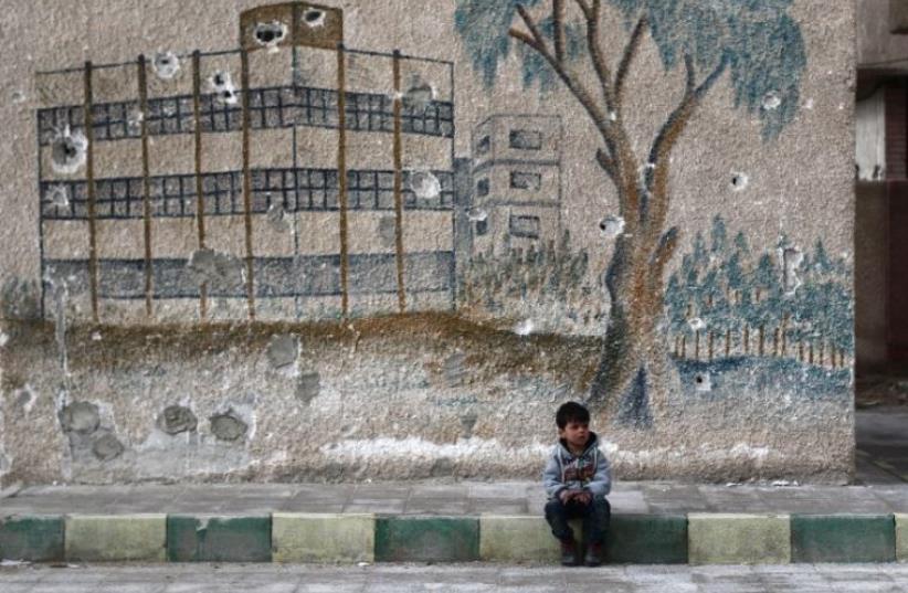 A Syrian child sits in front of a mural covered in bullet holes on the wall of a former school in the rebel-held region of Eastern Ghouta (photo credit: AFP PHOTO)