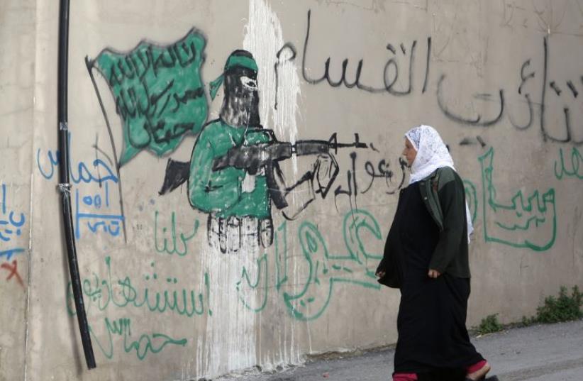 A Palestinian woman walks past a wall with graffiti depicting a gunman from the Al-Qassam brigades, the armed wing of the Hamas movement, in the West Bank village of Awarta.  The graffiti reads "All of us are martyrs." (photo credit: ABED OMAR QUSINI/REUTERS)