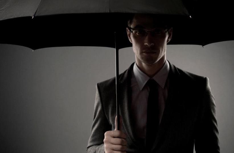 A mysterious man in a black suit standing in the shadows underneath an umbrella, looking like a spy or a secret agent [Illustrative] (photo credit: INGIMAGE)