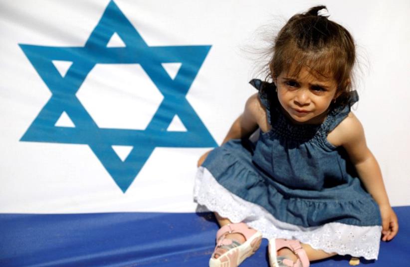 An Israeli girl sits on the national flag during a display of Israeli Defense Forces equipment and abilities, as part of the celebrations for Israel's Independence Day marking the 69th anniversary, in the southern city of Sderot, Israel May 2, 2017 (photo credit: REUTERS)