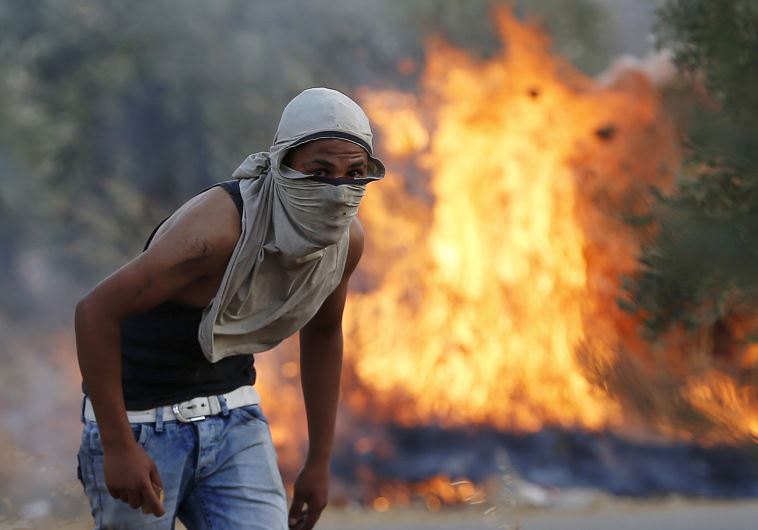 A Palestinian stone-thrower looks on as he stands in front of a fire during clashes with IDF troops in the West Bank village of Duma