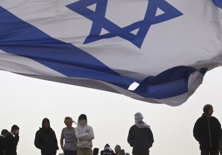An Israeli flag flutters near Israeli youths as they protest atop the roof of a synagogue in the Israeli settlement of Givat Ze'ev