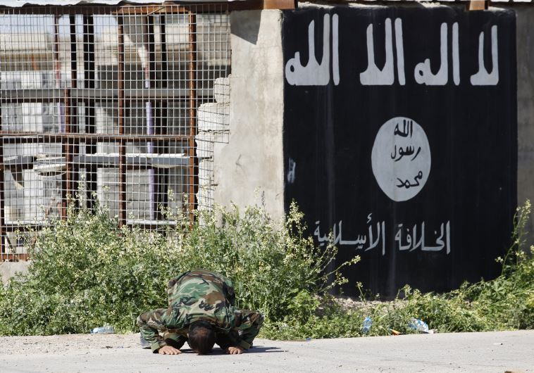 A member of a militia kneels as he celebrates victory next to a wall painted with the black flag commonly used by ISIS terrorists.