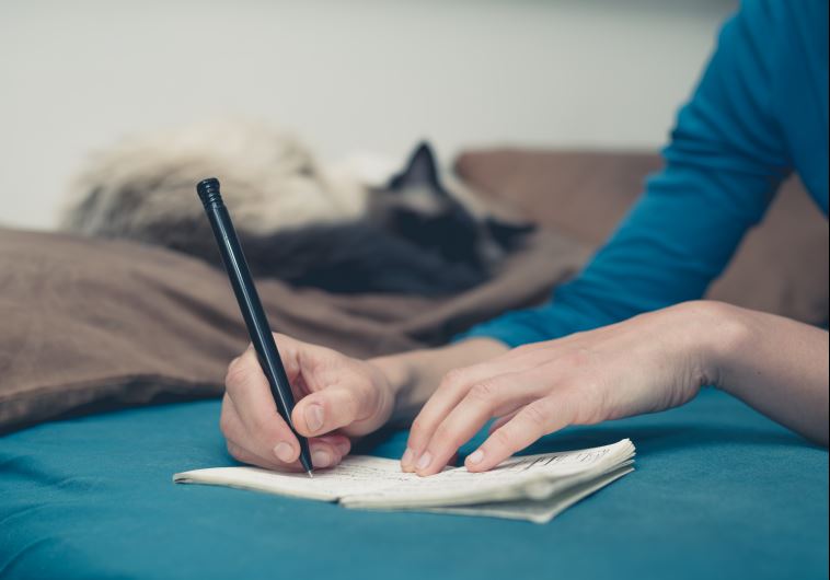 A young woman is lying in a bed with a cat and is taking notes with a pen and notepad