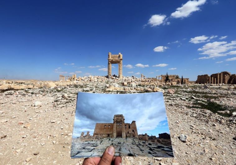A general view taken on March 31, 2016 shows a photographer holding his picture of the Temple of Bel taken on March 14, 2014 in front of the remains of the historic temple after it was destroyed by Islamic State