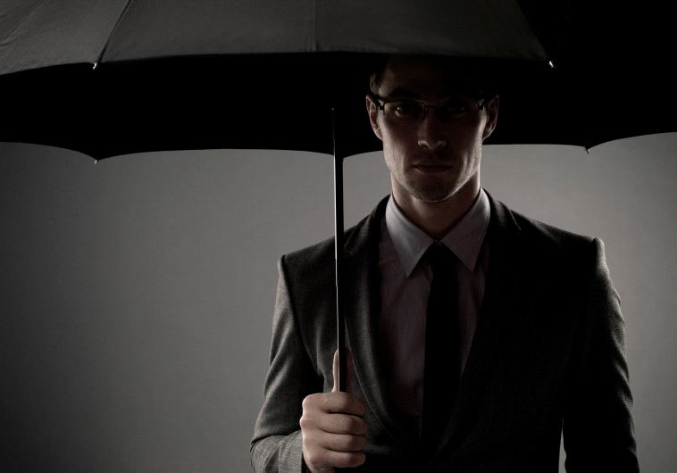 A mysterious man in a black suit standing in the shadows underneath an umbrella, looking like a spy or a secret agent [Illustrative]
